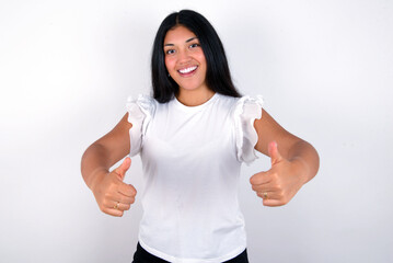 young brunette woman wearing white T-shirt standing against white background making positive gesture with thumbs up smiling and happy for success. Looking at the camera, winner gesture.