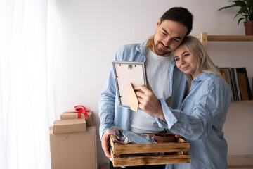 Young husband and wife moving to new place standing at home hugging holding frame looking at photo...
