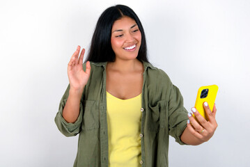 Portrait of happy friendly young brunette woman wearing green overshirt standing against white background taking selfie and waving hand, communicating on video call, online chatting.