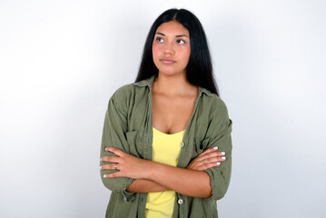 Charming thoughtful young brunette woman wearing green overshirt standing against white background stands with arms folded concentrated somewhere with pensive expression thinks what to do