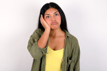 Sad lonely young brunette woman wearing green overshirt standing against white background touches cheek with hand bites lower lip and gazes with displeasure. Bad emotions