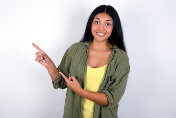 young brunette woman wearing green overshirt standing against white background points at copy space indicates for advertising gives right direction