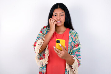 Portrait of pretty frightened young brunette woman wearing colourful outfit standing against white background chatting biting nails after reading some scary news on her smartphone.