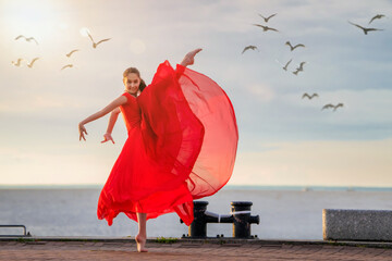 Dancing ballerina in a red flying skirt and leotard on the ocean embankment or on the sea beach surrounded by seagulls in the sky.