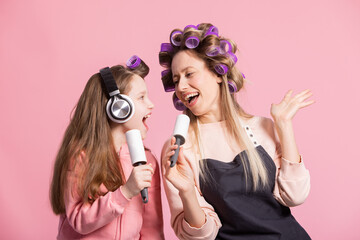 A homemade concert of a mother and daughter singing to clothes-cleaning rolls. Women have rollers to curl hair on head listen to music play on pink studio background.