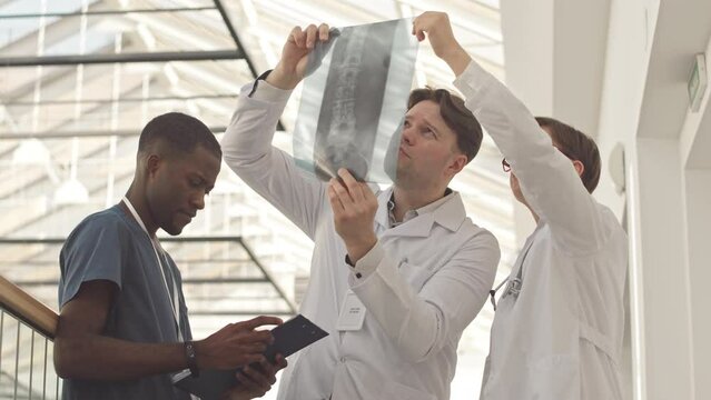 Medium slowmo of couple of doctors in lab coats examining patient xray image while their African American male assistant reading medical card of patient on clipboard, standing at clinic corridor