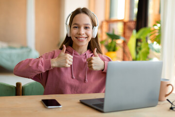 Distant education. Excited adolescent girl in headphones showing thumbs up and smiling while using laptop at home