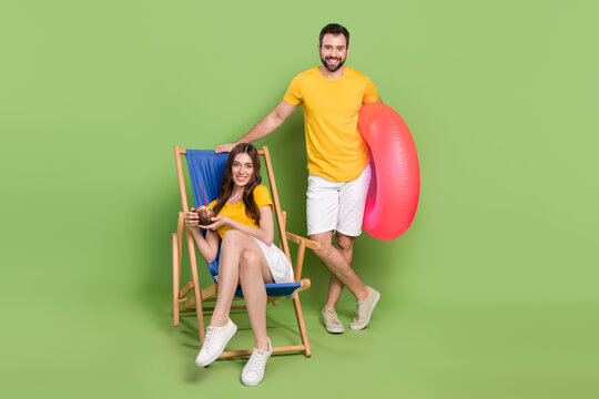 Full Size Portrait Of Two Peaceful Positive Partners Sitting Chaise Lounge Drink Cocktail Hold Inflatable Ring Isolated On Green Color Background