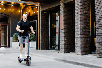 Young handsome man in white baseball cap riding on scooter to work along the street in the city