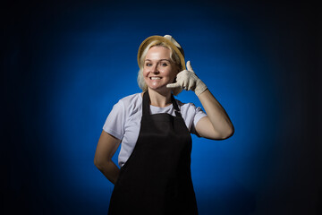 A woman in a white t-shirt, black apron, gloves and a hat shows a hand sign call me on a dark...
