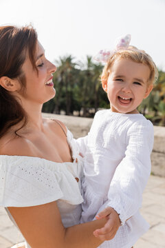 Portrait of baby girl in white dress looking at camera near mom in Valencia.