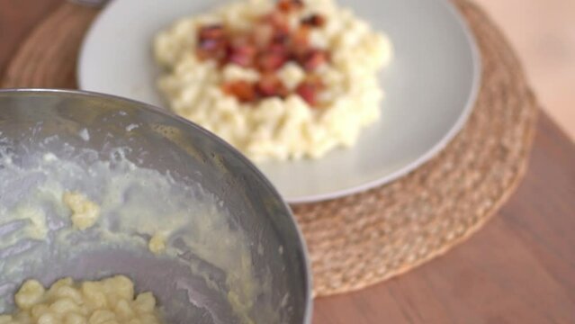 Bryndzove Halusky Dumpling In A Plate Topped With Cooked Bacon Bits, Prepared Potato Dough In A Mixing Bowl On The Side. close up, sliding rack focus