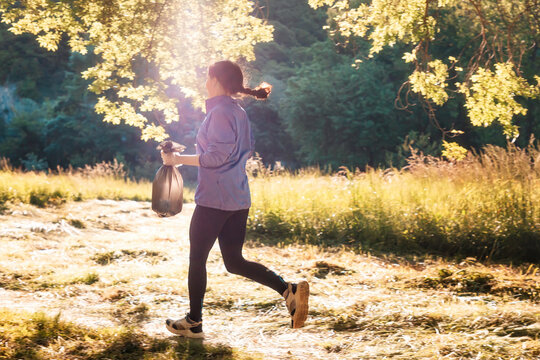 A young woman in sportswear runs through the park holding a bag of garbage in her hands. Back view. The concept of plogging and volunteering