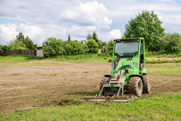 A green mini skid steer loader clear the construction site. Land work by the territory improvement. Small tractor with a ground leveler for moving soil, turf. Machine for agriculture work. Copy space - obrazy, fototapety, plakaty