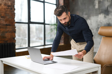 Busy businessman looking through the workpaper and using laptop in modern loft office, hurrying to finish report in time