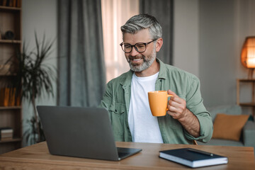 Cheerful old caucasian man with beard in glasses typing on laptop at workplace, drinking favorite...