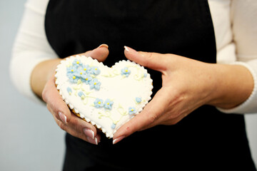 A woman in a black apron holds a heart-shaped gingerbread with painted flowers in her hands