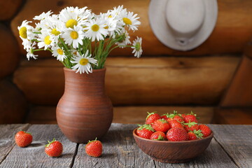 Obraz na płótnie Canvas A clay bowl of ripe strawberries next to a bouquet of field daisies in a pottery vase against a log wall with a straw hat hanging on it.