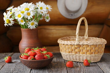 A clay bowl of ripe strawberries next to a bouquet of field daisies in a pottery vase against a log wall with a straw hat hanging on it.