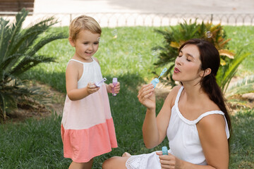 Positive kid holding soap bubbles near mother on lawn.