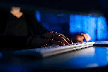 Close-up hands of unrecognizable hacker man working typing on keyboard laptop computer sitting at desk in dark room with blue neon lights. Concept of cyber attack, virus, malware, illegally. - Powered by Adobe