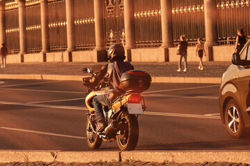 A motorcyclist travels on a motorcycle around the city. A man rides among cars on a city road in the sun at sunset.