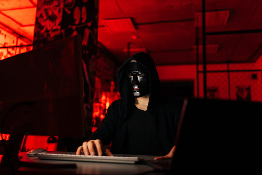 Dangerous unrecognizable hacker man wearing in black mask and sweatshirt hacking online web site using laptop and desktop, sitting at table in dark basement room with red neon lights.