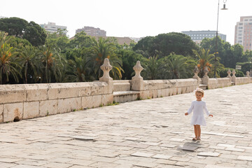Smiling toddler girl in white summer dress walking on Puente Del Mar bridge in Valencia.
