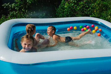 Three happy friends are splashing in an inflatable pool in the garden. Concept of summer season and recreation. Kids having fun together. Children playing and swimming in water on the backyard.