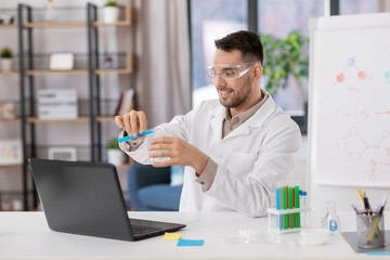 distance education, school and science concept - happy smiling male chemistry teacher in goggles with laptop computer having online class and pouring chemical from test tube to flask at home office