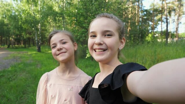 Couple of teen girl friends makes selfie on phone grimacing. School vloggers wearing stylish dresses make vlogs smiling and enjoying walking on park lawn