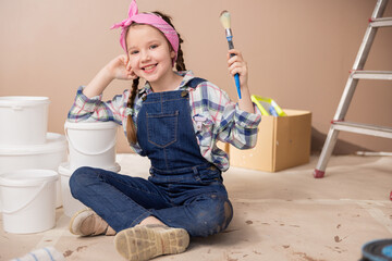 Joyful child in denim clothes soiled with brown wall paint sits on the floor holding a paintbrush...