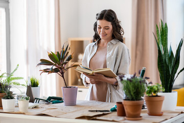 people, gardening and housework concept - happy smiling woman with book planting pot flowers at home