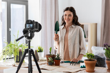 blogging, gardening and housework concept - happy woman or blogger with camera and trowel planting pot flowers and recording tutorial video at home