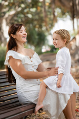 Cheerful toddler child in white dress sitting on mother in summer park.