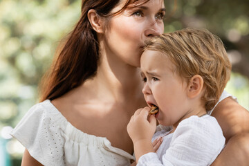 Fototapeta premium Woman kissing daughter with cookie in summer park.