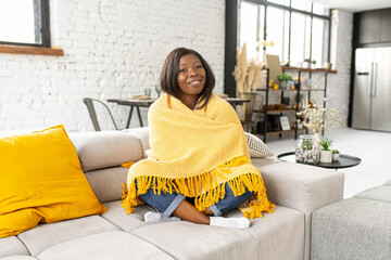 Serene African american woman enjoying weekend at home, sitting on the sofa wrapped with blanket and laughing, calm lady resting, looks at the camera and smiles