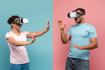 Young black spouses in VR headset touching air during virtual reality experience, blue and pink studio background