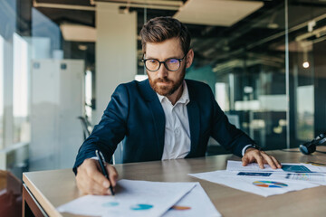 Concentrated handsome businessman in glasses working with documents in office interior, checking and signing papers