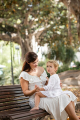 Woman in summer dress holding hand of baby daughter on bench in park.