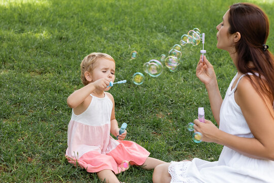 Daughter in dress blowing soap bubbles near mom on lawn in park.