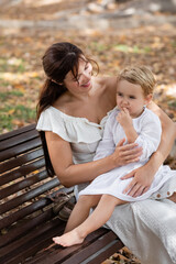 Smiling woman in dress hugging toddler daughter on bench in park.