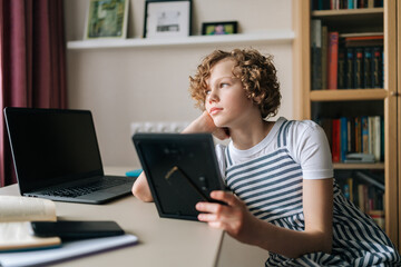 Missed preteen girl holding photo frame with family portrait in hands and looking out window with...