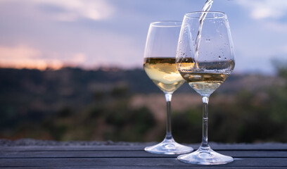 Two elegant glasses of white wine on a table with countryside in the background, at sunset