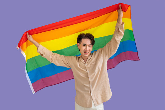 Portrait of cute Asian LGBT homosexual men or gay standing and smiling, holding pride rainbow flag on purple background. Looking at camera.