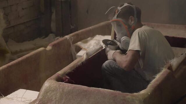 A worker wearing a protective mask is working with an angle grinder. A man is cutting off excess parts on fiberglass hull of a boat. Slow motion. Small-scale production of boats in a private workshop