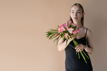 young woman in black strap dress holding bouquet of pink flowers isolated on beige.