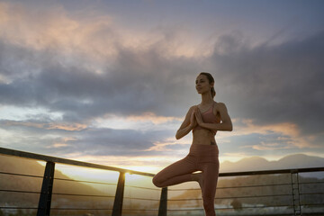 Young sporty woman practicing yoga outdoor standing in Vrksasana exercise, Tree pose, working out. Beautiful dark sunset sky and mountains landscape on background
