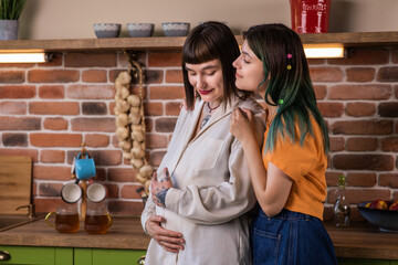 Good looking lesbian couple very enthusiastic standing beside the country style kitchen they waiting a baby young lady partner caresses the belly of her pregnant girlfriend concept of LGBT freedom