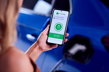 Woman Charging Electric Car With Cable Using App On Phone To Monitor Battery Level